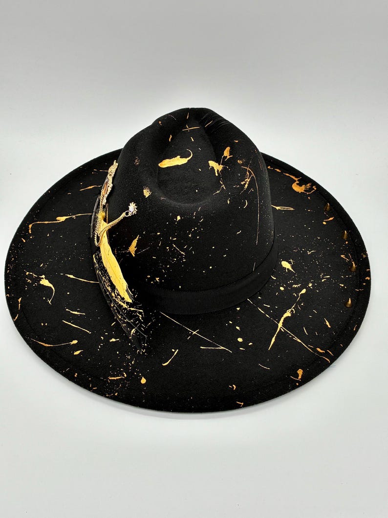 Op de afbeelding: Een zwarte fedora hoed met een brede rand en een ingedeukte kroon, versierd met gouden verfspatten over het oppervlak. Een zwarte grosgrain lintband omcirkelt de basis van de kroon, geaccentueerd door een goudkleurige veer en metalen details.