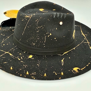 Op de afbeelding: Een zwarte fedora hoed met een goudkleurig verfspatpatroon over de kroon en de brede rand. De hoed is voorzien van een zwarte lintband, vier kleine zilverkleurige metalen studs aan de zijkant, een witte knop en een gele veer als accent.
