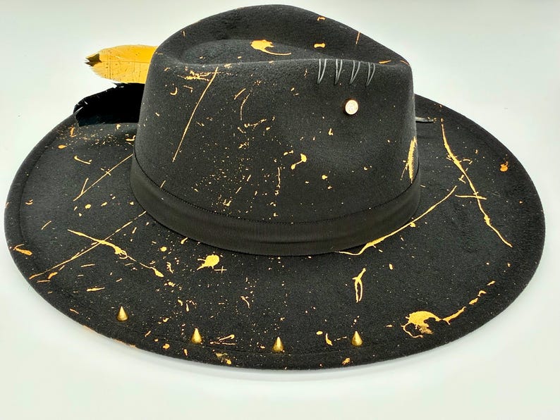 Op de afbeelding: Een zwarte fedora hoed met een goudkleurig verfspatpatroon over de kroon en de brede rand. De hoed is voorzien van een zwarte lintband, vier kleine zilverkleurige metalen studs aan de zijkant, een witte knop en een gele veer als accent.