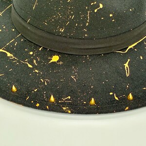 Op de afbeelding: Een zwarte hoed met een brede rand en een zwarte grosgrain lintband rond de kroon. Het oppervlak is versierd met een geel verfspatpatroon en een reeks kleine, goudkleurige, conische metalen studs langs de rand van de hoed.