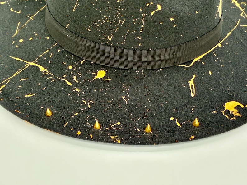 Op de afbeelding: Een zwarte hoed met een brede rand en een zwarte grosgrain lintband rond de kroon. Het oppervlak is versierd met een geel verfspatpatroon en een reeks kleine, goudkleurige, conische metalen studs langs de rand van de hoed.