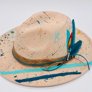 May include: A beige fedora hat with a splattered paint design in shades of blue and black. The hat has a brown rope band, a turquoise band, and a decorative feather accent with blue, red, and yellow feathers.