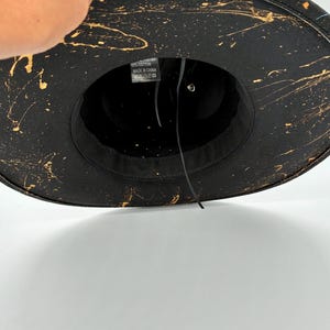 Op de afbeelding: Een zwarte fedora hoed met een goudkleurig verfspatpatroon over de rand en de kroon. De binnenkant is voorzien van een zwarte stoffen zweetband en een wit waslabel met de tekst 65% katoen, Made in China en diverse wassymbolen.