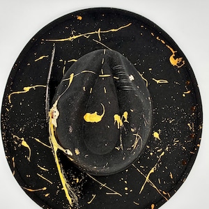 Op de afbeelding: Een zwarte hoed met een brede rand, een ingedeukte kroon en gouden verfspatten over het oppervlak. De hoed is versierd met een donkere veer, een metalen decoratief element met een kleine rode steen en zwarte koorddetails op de kroon.