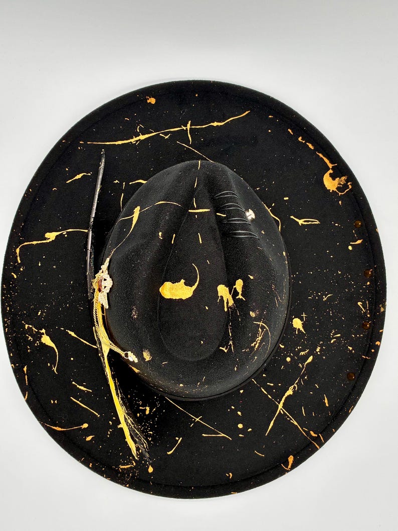 Op de afbeelding: Een zwarte hoed met een brede rand, een ingedeukte kroon en gouden verfspatten over het oppervlak. De hoed is versierd met een donkere veer, een metalen decoratief element met een kleine rode steen en zwarte koorddetails op de kroon.