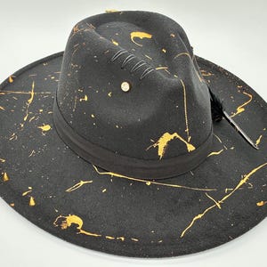 Op de afbeelding: Een zwarte fedora hoed met een brede rand en een ingedeukte kroon. De hoed is versierd met gouden verfspatten over het oppervlak, een zwarte lintband, een kleine metalen stud en een enkele zwarte veer die aan de zijkant is ingestoken.