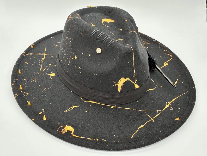 Op de afbeelding: Een zwarte fedora hoed met een brede rand en een ingedeukte kroon. De hoed is versierd met gouden verfspatten over het oppervlak, een zwarte lintband, een kleine metalen stud en een enkele zwarte veer die aan de zijkant is ingestoken.