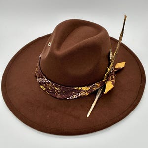 Può includere: Un cappello fedora marrone scuro con tesa larga e corona pizzicata. Il cappello è decorato con una fascia a bandana marrone scuro con un motivo paisley giallo e bianco, annodata sul lato con una piuma lunga, sottile e maculata.