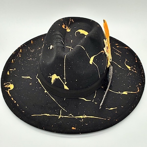 Op de afbeelding: Een zwarte fedora hoed met een brede rand en een ingedeukte kroon. De hoed is versierd met gele verfspatten over het oppervlak, een zwarte lintband, een reeks zwarte stiksels en een decoratief accent in de vorm van een veer.