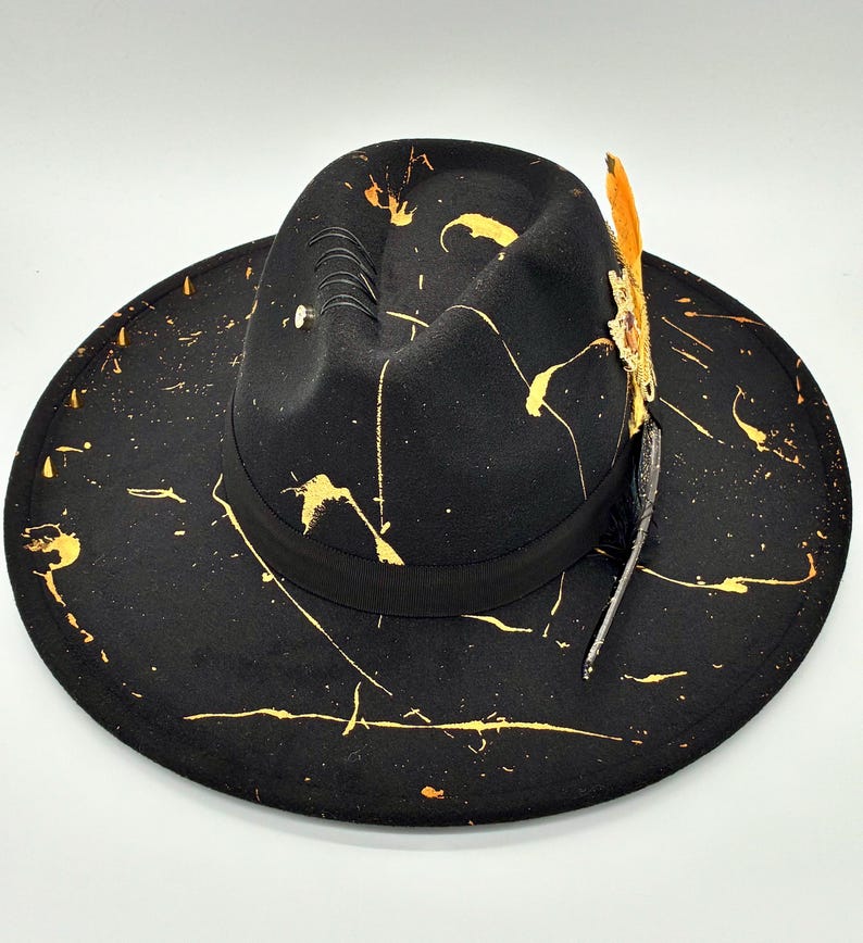 Op de afbeelding: Een zwarte fedora hoed met een brede rand en een ingedeukte kroon. De hoed is versierd met gele verfspatten over het oppervlak, een zwarte lintband, een reeks zwarte stiksels en een decoratief accent in de vorm van een veer.