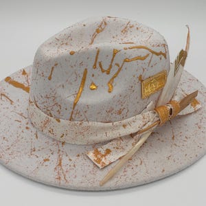 May include: A white fedora hat with gold paint splatter design. The hat features a gold band, a decorative gold emblem, and feathers. The hat's brim is wide, and the overall style is artistic and fashionable.