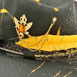Op de afbeelding: Een zwarte fedora hoed met een geel verfspatpatroon, een zwarte lintband en een gele veer. Een goudkleurige broche in de vorm van een gevleugelde kroon met een amberkleurige rechthoekige steen is bevestigd aan een gouden ketting met een zonnetje.