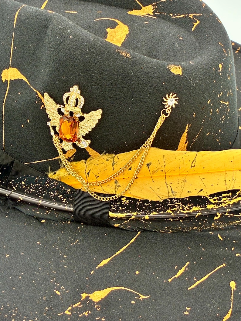 Op de afbeelding: Een zwarte fedora hoed met een geel verfspatpatroon, een zwarte lintband en een gele veer. Een goudkleurige broche in de vorm van een gevleugelde kroon met een amberkleurige rechthoekige steen is bevestigd aan een gouden ketting met een zonnetje.