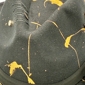 Op de afbeelding: Een zwarte vilten fedora hoed met een levendig oranje verfspatpatroon over de kroon. De hoed is voorzien van een zwarte grosgrain lintband, een kleine metalen stud en vier parallelle zwarte decoratieve stiksels op de zijkant van de kroon.