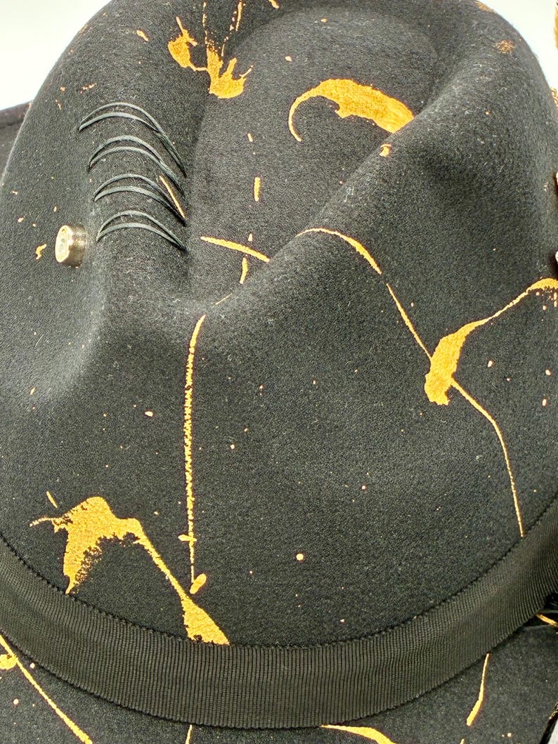 Op de afbeelding: Een zwarte vilten fedora hoed met een levendig oranje verfspatpatroon over de kroon. De hoed is voorzien van een zwarte grosgrain lintband, een kleine metalen stud en vier parallelle zwarte decoratieve stiksels op de zijkant van de kroon.