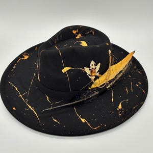 May include: A black fedora hat featuring a gold paint splatter pattern across the crown and wide brim. The hat is adorned with a black ribbon band, a gold-toned crown emblem with an amber-colored stone, a gold chain, and a long, dark feather.