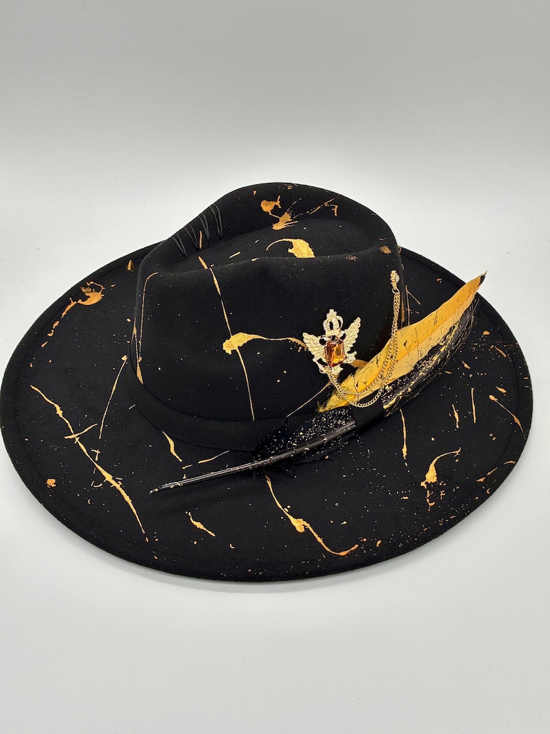 Op de afbeelding: Een zwarte fedora hoed met een goudkleurig verfspatpatroon over de kroon en de brede rand. De hoed is versierd met een zwarte lintband, een goudkleurig kroonembleem met een barnsteenkleurige steen, een gouden ketting en een lange, donkere veer.