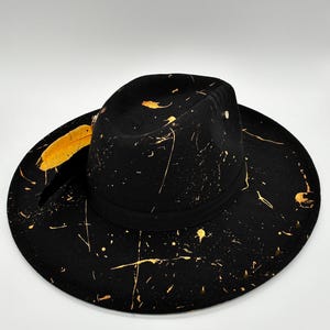 Op de afbeelding: Een zwarte fedora hoed met een brede rand en een gestructureerde kroon, versierd met een goudkleurig verfspatpatroon over het oppervlak. Een zwarte band omsluit de basis van de kroon en een gele veer is aan de zijkant ingestoken.