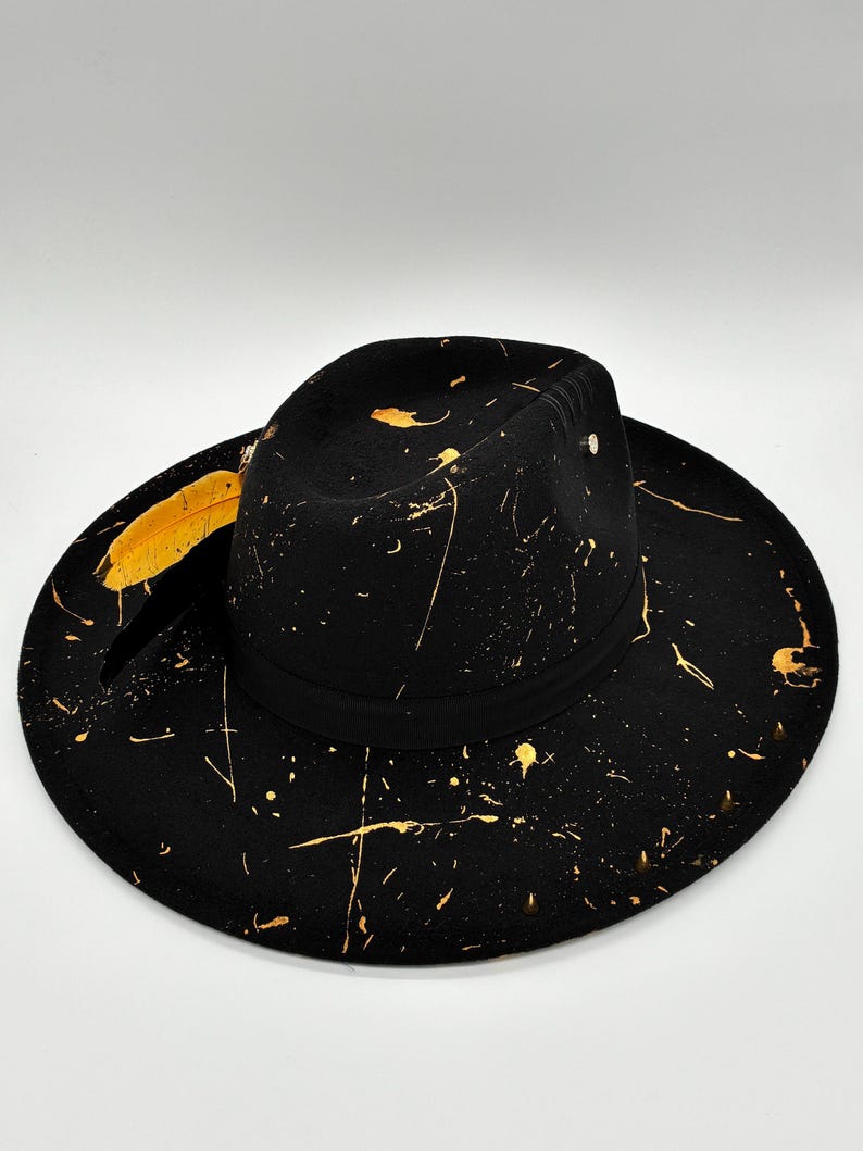 Op de afbeelding: Een zwarte fedora hoed met een brede rand en een gestructureerde kroon, versierd met een goudkleurig verfspatpatroon over het oppervlak. Een zwarte band omsluit de basis van de kroon en een gele veer is aan de zijkant ingestoken.