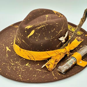 Puede incluir: Un sombrero fedora marrón con un patrón de salpicaduras de pintura amarilla, una banda de tela amarilla, un broche de pájaro plateado y una pluma oscura larga. Un tubo de cigarro de plástico transparente con funda de cuero amarilla descansa en el ala.