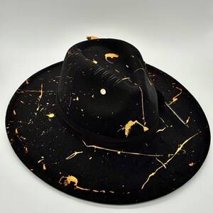 Op de afbeelding: Een zwarte fedora hoed met een brede rand en een ingedeukte kroon. De hoed is versierd met een gouden verfspatpatroon over het oppervlak. Een zwarte lintband omsluit de basis van de kroon en een kleine metalen stud is zichtbaar op de zijkant.