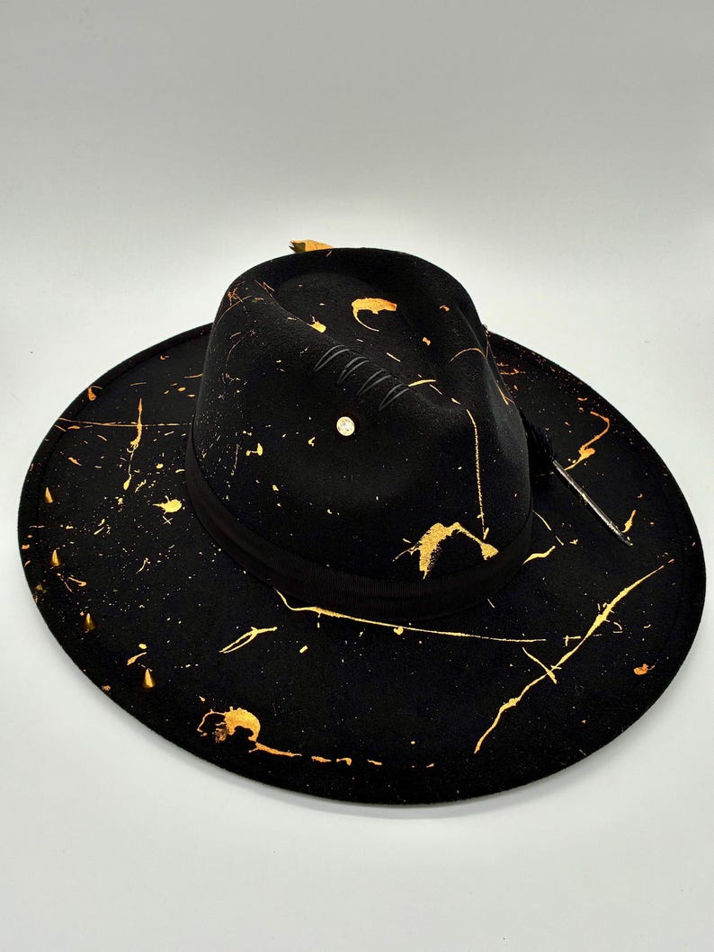 Op de afbeelding: Een zwarte fedora hoed met een brede rand en een ingedeukte kroon. De hoed is versierd met een gouden verfspatpatroon over het oppervlak. Een zwarte lintband omsluit de basis van de kroon en een kleine metalen stud is zichtbaar op de zijkant.