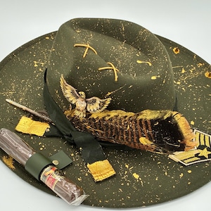 Puede incluir: Un sombrero fedora verde oliva con salpicaduras de pintura dorada, una cinta negra, un broche de águila dorada, una pluma rayada y un parche militar. Un cigarro en un estuche de cuero verde está colocado sobre el ala del sombrero.