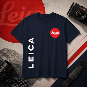 Puede incluir: Una camiseta de manga corta azul marino que presenta un logotipo circular rojo con el texto blanco Leica en la parte superior derecha del pecho y la palabra LEICA impresa verticalmente en letras mayúsculas blancas en el lado izquierdo.