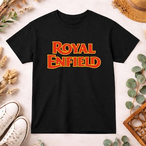 May include: A black short-sleeved t-shirt featuring the text ROYAL ENFIELD printed in a bold, stylized yellow font with a red outline centered on the chest. The garment has a classic crew neckline and a straight hemline.