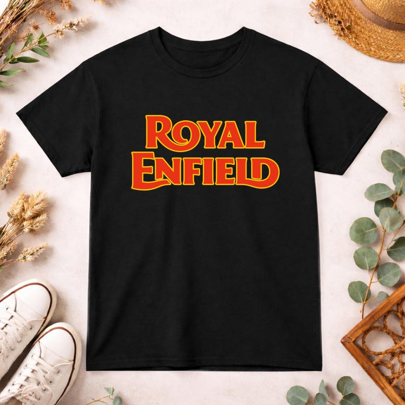 May include: A black short-sleeved t-shirt featuring the text ROYAL ENFIELD printed in a bold, stylized yellow font with a red outline centered on the chest. The garment has a classic crew neckline and a straight hemline.