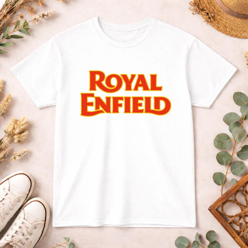 May include: A white short-sleeved t-shirt featuring the text ROYAL ENFIELD printed in a bold, stylized red font with a thin yellow outline centered on the chest. The garment has a classic crew neckline and a standard straight-cut silhouette.