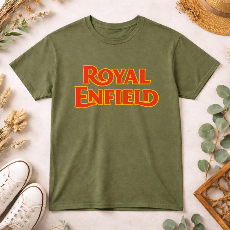 May include: An olive green short-sleeve t-shirt featuring the text ROYAL ENFIELD printed in a bold, stylized red font with a yellow outline centered on the chest. The garment has a classic crew neckline and a standard straight-cut hemline.