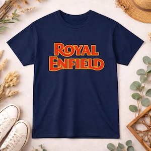 May include: A navy blue short-sleeve t-shirt featuring the text ROYAL ENFIELD printed in a bold, stylized yellow font with a red outline centered on the chest. The garment has a classic crew neckline and a straight hemline design.