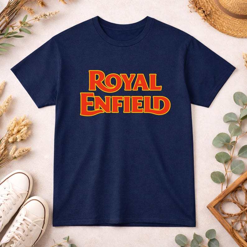 May include: A navy blue short-sleeve t-shirt featuring the text ROYAL ENFIELD printed in a bold, stylized yellow font with a red outline centered on the chest. The garment has a classic crew neckline and a straight hemline design.