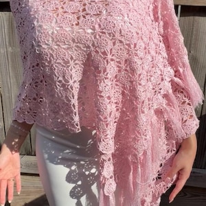 May include: A light pink, open-weave crochet shawl with a floral pattern. The shawl has a ruffled collar and fringe detailing along the edges. The garment is worn over a white dress.