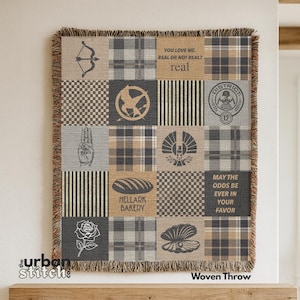 May include: Woven throw blanket with a patchwork design featuring various symbols and text from a popular book series. The blanket has a fringed edge and includes phrases like "You love me. Real or not real? real" and "May the odds be ever in your favour."