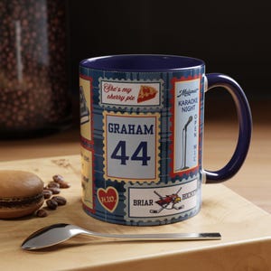 Peut inclure: Un mug en céramique bleu foncé avec un fond à carreaux bleus et divers graphismes rectangulaires en forme de timbres-poste. Le graphisme central affiche le texte "GRAHAM 44" en lettres bleues grasses. Les autres motifs incluent "She's my cherry pie", "Maloney's KARAOKE NIGHT", un microphone, un cœur avec "H.W." et un logo "BRIAR HOCKEY".