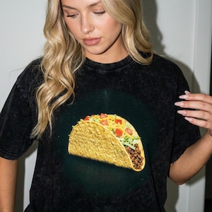 Könnte beinhalten: Schwarzes T-Shirt mit einem großen, farbenfrohen Taco-Motiv. Der Taco ist mit gewürztem Hackfleisch, gerassem Salat, gewürfelten Tomaten und geriebenem Käse gefüllt. Das Shirt hat einen Vintage-Look.