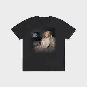 May include: Black t-shirt featuring a doll in a bathrobe holding a glass of wine, watching TV. The doll has blonde hair and is seated on a floral patterned couch. The TV screen shows a blurry image.