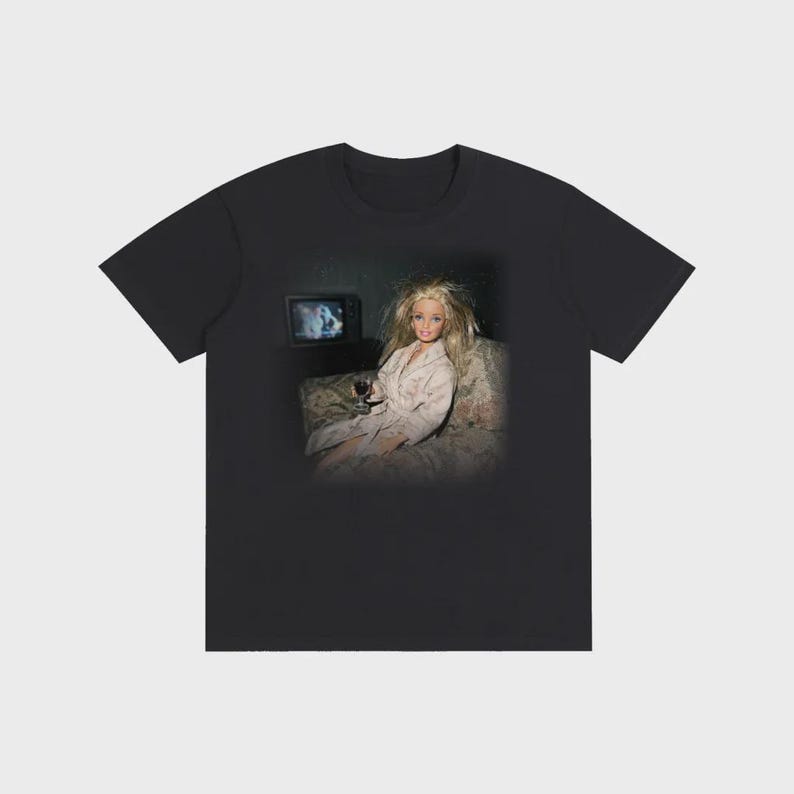 May include: Black t-shirt featuring a doll in a bathrobe holding a glass of wine, watching TV. The doll has blonde hair and is seated on a floral patterned couch. The TV screen shows a blurry image.