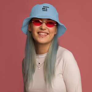 May include: A light blue bucket hat with the text "Business BIG Album" in black. The person is wearing red-tinted sunglasses with a gold frame and a white long-sleeve shirt. The background is a solid pink color.