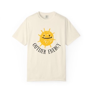 May include: Cream-colored t-shirt featuring a yellow sun graphic with a smiling face and the text "OUTLIER ENERGY" in black. The shirt is a casual, short-sleeved design, perfect for everyday wear. The graphic has a hand-drawn aesthetic.
