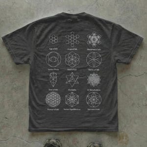 Peut inclure: T-shirt gris foncé orné de motifs géométriques blancs, dont l'Œuf de Vie, le Fruit de Vie et le Cube de Métatron. Le t-shirt présente divers symboles de géométrie sacrée, idéal pour les passionnés de spiritualité et d'ésotérisme.