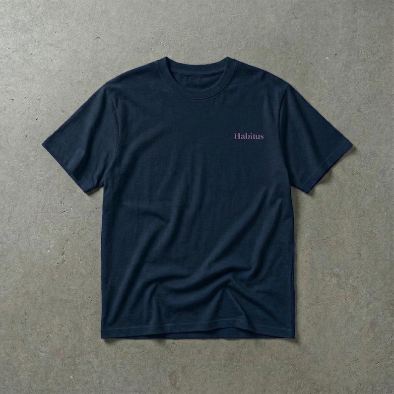 May include: Navy blue t-shirt with the word "Habitus" printed in a pinkish-purple color on the upper right chest area. The shirt is made of a soft material and has short sleeves and a crew neck.
