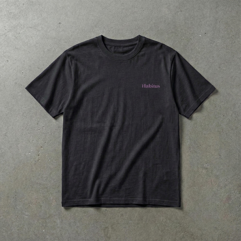 May include: A dark gray t-shirt with short sleeves. The word "Habitus" is printed in a small, purple font on the upper chest area. The shirt is made of a soft material and is suitable for casual wear.