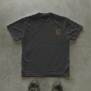 May include: A dark gray t-shirt with a crew neck and short sleeves. A small, gold-colored square graphic with the word "Habitat" is printed on the upper left chest area. The shirt is lying on a concrete surface.