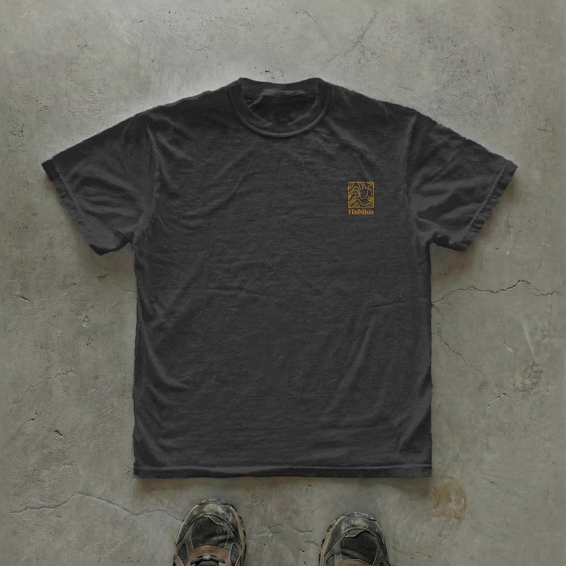 May include: A dark gray t-shirt with a crew neck and short sleeves. A small, gold-colored square graphic with the word "Habitat" is printed on the upper left chest area. The shirt is lying on a concrete surface.