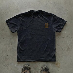 May include: Black t-shirt with a small gold-colored square graphic on the upper left chest. The graphic features an abstract design and the word "Habitus" below it. The shirt is made of a soft material and has short sleeves.