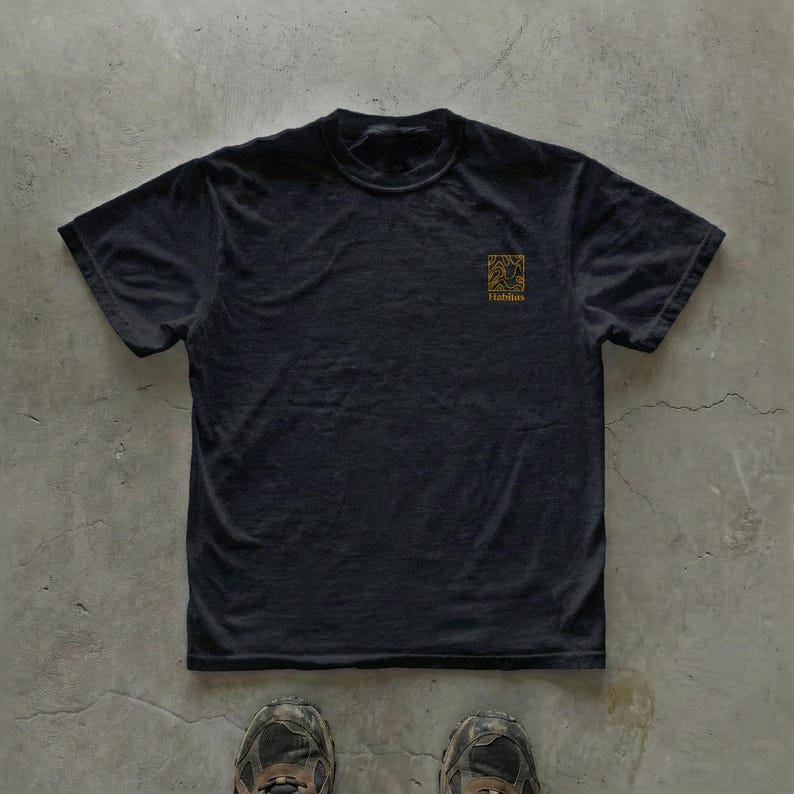May include: Black t-shirt with a small gold-colored square graphic on the upper left chest. The graphic features an abstract design and the word "Habitus" below it. The shirt is made of a soft material and has short sleeves.