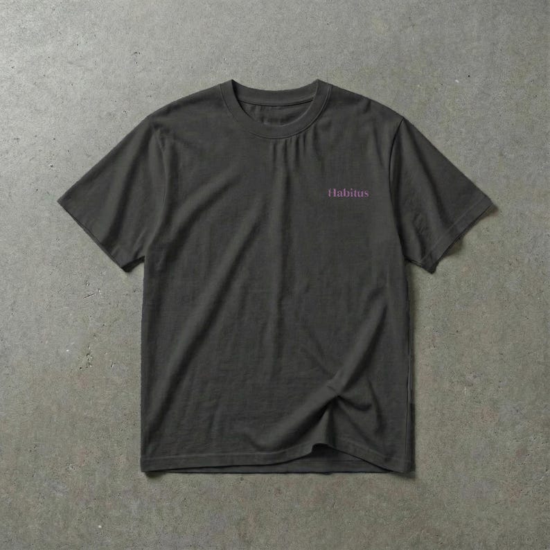 May include: A dark gray t-shirt with short sleeves. The word "Habitus" is printed in a light purple color on the upper left chest area of the shirt. The shirt is made of a soft, comfortable material.